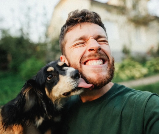 A man smiles for a selfie while his dog playfully licks his face. A man smiles for a selfie while his dog playfully licks his face.
